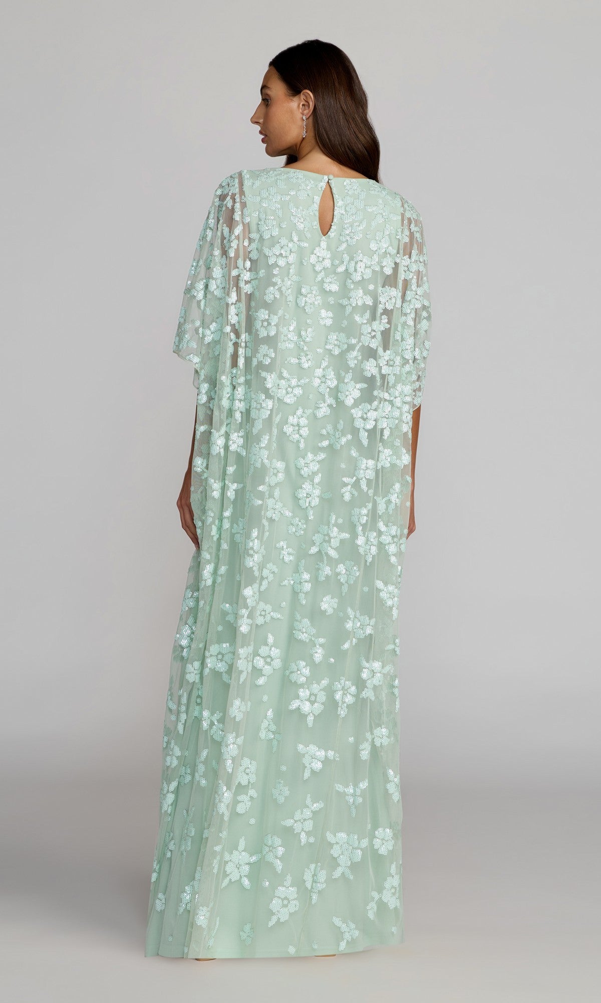 A woman is seen from the back wearing the Mac Duggal 9441 Long Formal Dress, a floor-length, light green evening gown adorned with intricate sequined floral patterns on a sheer overlay, featuring cape sleeves and a keyhole cutout at the neckline.